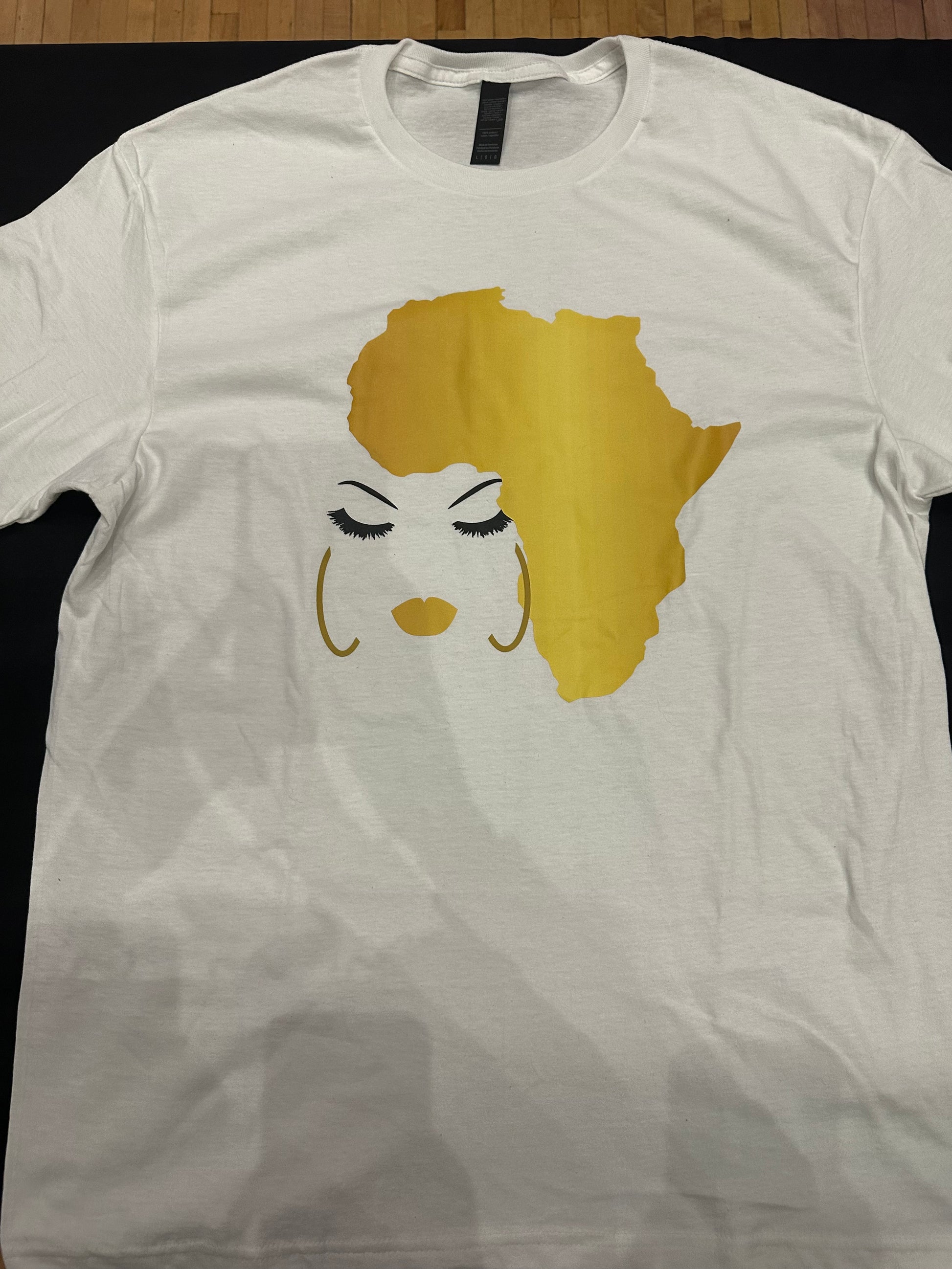 White shirt with Africa in a yellow golden hue with black eyebrows and eyelashes with golden hue lips and gold earrings. Africa is used as her hair. Melanin apparel.