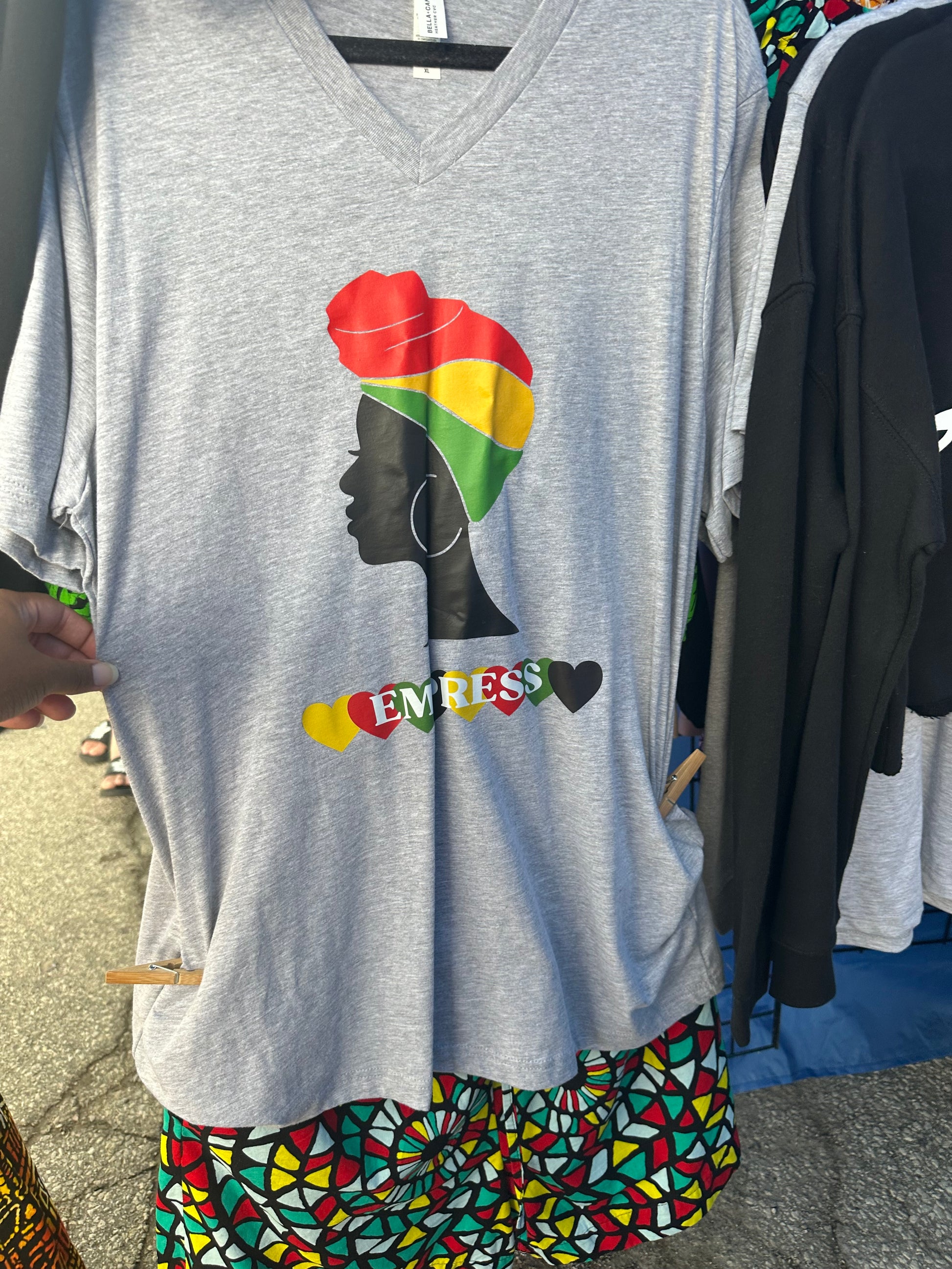 Heather Grey V-neck shirt with black woman silhouette with red, yellow and green headwrap. Below the silhouette hearts yellow, red, green and black repeatedly with word Empress in white. Melanin apparel.