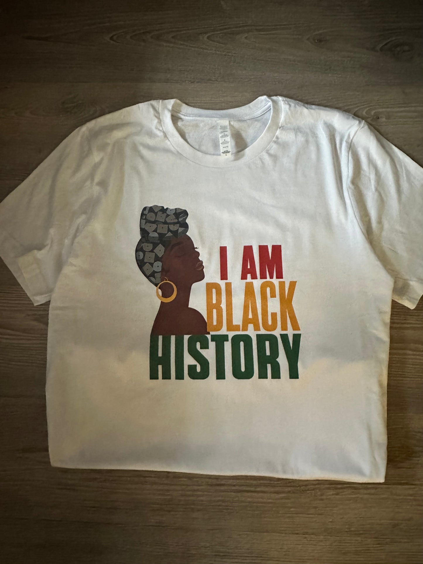 I am black history white tshirt round neck with a dark brown skin tone woman wearing different shades of blue headwrap with gold earring with text I am in red Black in yellow History in green all caps. Melanin Apparel. I am black history.