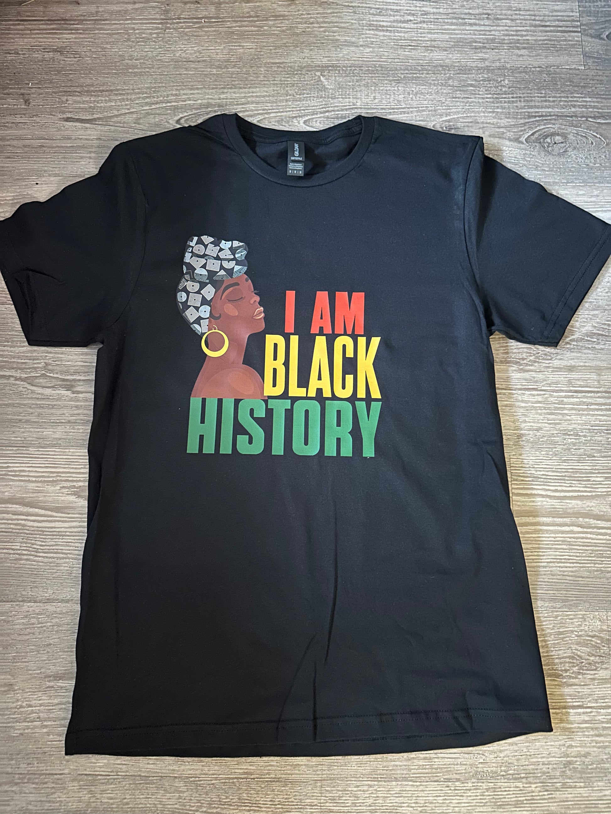 I am black history black tshirt round neck with a dark brown skin tone woman wearing different shades of blue headwrap with gold earring with text I am in red Black in yellow History in green all caps. Melanin Apparel. I am black history.