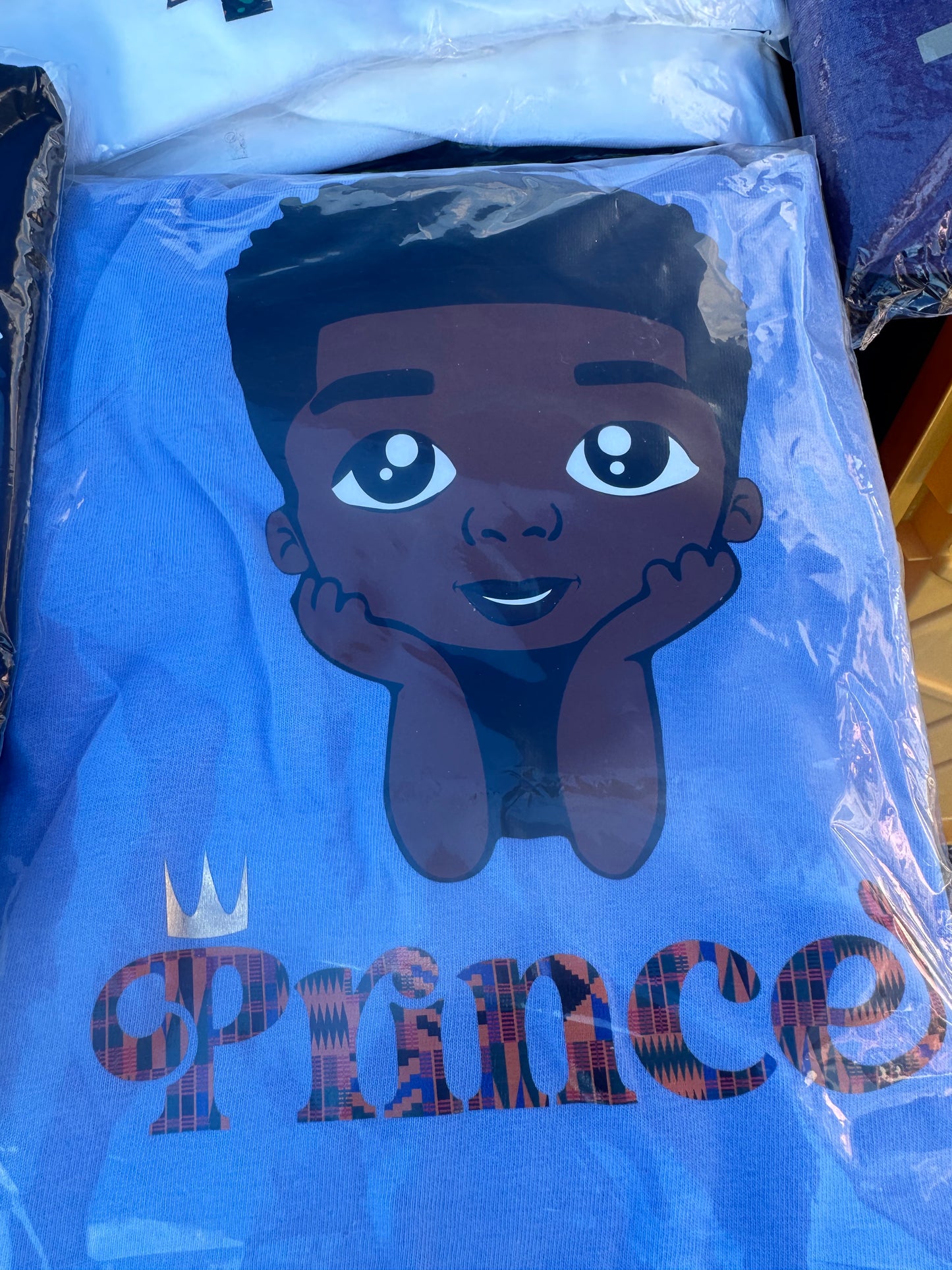 Blue shirt with black boy with hands under chin with word Prince in African print and gold crown over the P. Melanin apparel.