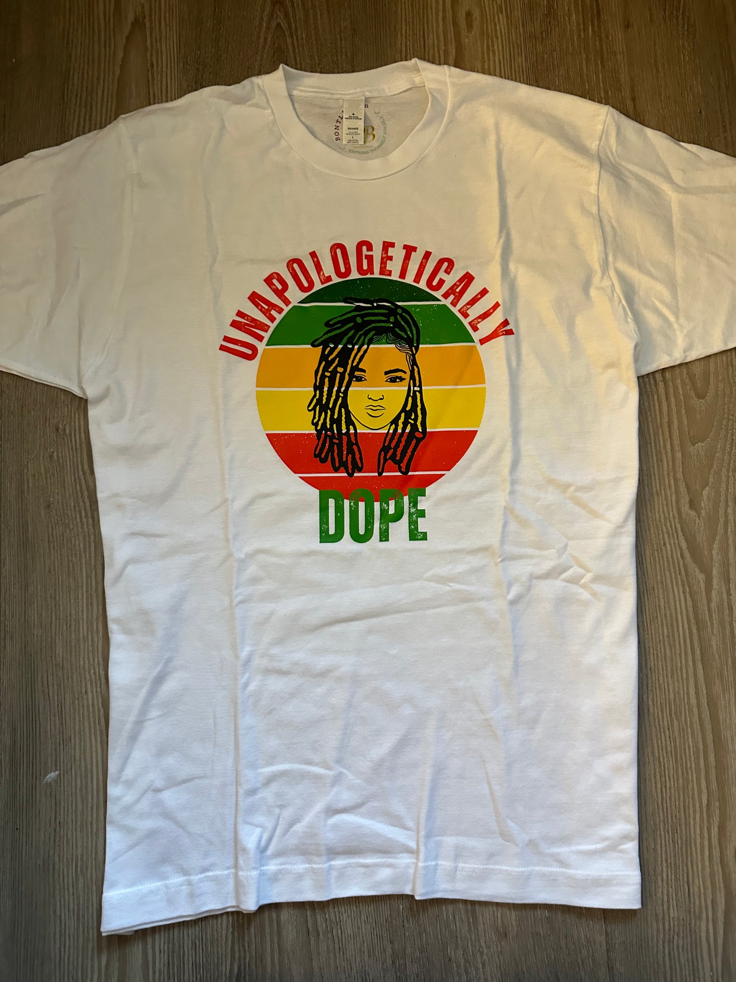 Unapologetically in Red text Dope in green text with green, orange, yellow, red retro circle with dreadlocks lady on top on a white shirt. Melanin Apparel. 