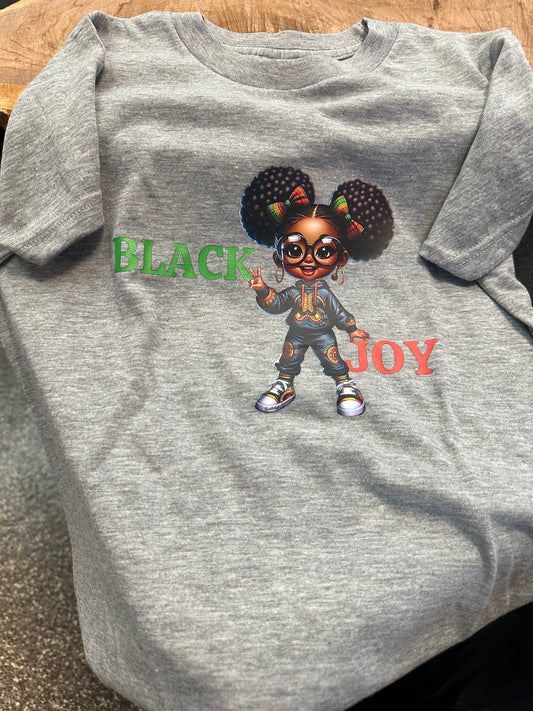 Grey shirt with green text Black with a black girl with two puffs, glasses wearing African print jumpsuit and African print kicks in the middle with word Joy in red. Melanin apparel. Black girl joy