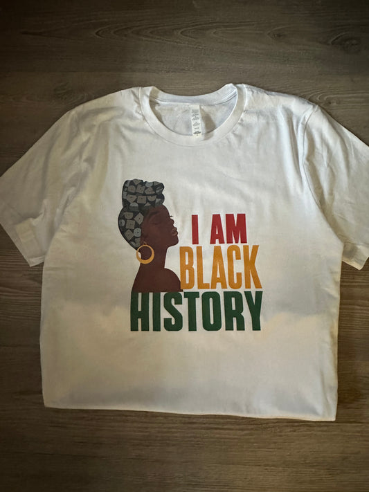 I am black history white tshirt round neck with a dark brown skin tone woman wearing different shades of blue headwrap with gold earring with text I am in red Black in yellow History in green all caps. Melanin Apparel. I am black history. 