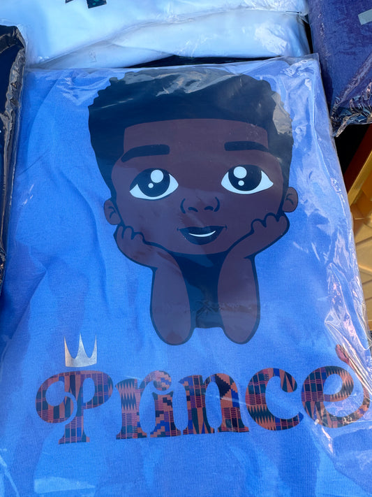 Blue shirt with black boy with hands under chin with word Prince in African print and gold crown over the P. Melanin apparel. 